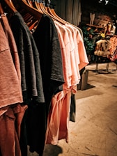 A row of t-shirts in varying shades of pink and black hangs neatly on wooden hangers. The background features a retail space with dim lighting, rustic wooden walls, and some decorative plants and clothing displays.