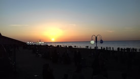 The scene captures a beautiful sunset over the ocean with a warm orange and yellow sky. A crowd of people is gathered along the shoreline, silhouetted against the evening light. The image also features street lamps with a unique curved design, and the atmosphere suggests a leisurely evening by the beach.