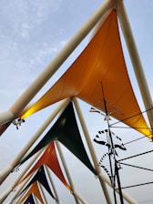 Triangular fabric structures are arranged in a pattern, attached to tall, white, triangular frameworks. Each fabric is a different color, primarily orange, green, and grey. A series of black and white abstract shapes hang on rods, adding an artistic element. The sky in the background is overcast with light cloud cover.