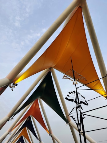 Triangular fabric structures are arranged in a pattern, attached to tall, white, triangular frameworks. Each fabric is a different color, primarily orange, green, and grey. A series of black and white abstract shapes hang on rods, adding an artistic element. The sky in the background is overcast with light cloud cover.