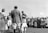 A group of people is gathered in an outdoor setting on a sunny day. In the foreground, individuals dressed in equestrian or formal attire are standing around a table covered with a cloth, possibly engaging in a ceremony or award presentation. Behind them, a crowd of onlookers, including men, women, and children in mid-20th-century clothing, observes the event.