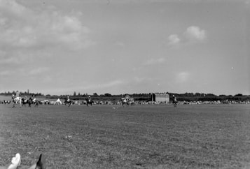 People riding horses across a large open field with spectators in the distance. A small group of tents and a notable structure appear along the horizon under a partly cloudy sky.
