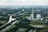A large industrial facility with tall chimneys is located near a river surrounded by greenery and farmland. The landscape is expansive, with roads and bridges crossing the area. Small residential and industrial buildings are visible in the distance.