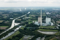 A large industrial facility with tall chimneys is located near a river surrounded by greenery and farmland. The landscape is expansive, with roads and bridges crossing the area. Small residential and industrial buildings are visible in the distance.