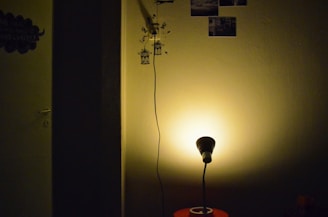 A dimly lit room featuring a small lamp casting a warm glow on a plain wall. The corner of the room is partially visible with wall decorations such as small photo prints and a decal of birds near a hanging cord. A dark shadow divides part of the scene, adding contrast to the illuminated section.