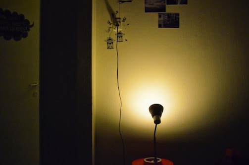 A dimly lit room featuring a small lamp casting a warm glow on a plain wall. The corner of the room is partially visible with wall decorations such as small photo prints and a decal of birds near a hanging cord. A dark shadow divides part of the scene, adding contrast to the illuminated section.