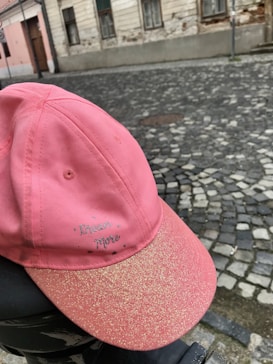 A pink baseball cap with the words 'Dream More' embroidered on it is placed on a black surface. The cap has a sparkly, glittery appearance on the brim. The background features a cobblestone street and an old, weathered building with peeling paint and mismatched windows.