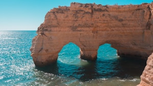 A natural rock formation with two large arches stands prominently against a backdrop of clear turquoise waters. The sunlight casts gentle reflections on the water's surface, creating a serene and peaceful atmosphere. The rock has a rugged, textured appearance, adding to the scenic beauty of the coastal landscape.