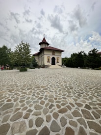 A small church with a red-roofed tower and decorative icons on the front stands amid a paved courtyard. Green trees frame the scene on both sides under a cloudy sky.