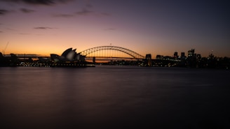 A panoramic view of a cityscape at dusk, featuring a famous opera house with unique architectural design beside a large arch bridge spanning across a body of water. The skyline is faintly illuminated against the darkening sky.