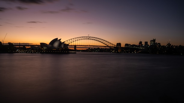 A panoramic view of a cityscape at dusk, featuring a famous opera house with unique architectural design beside a large arch bridge spanning across a body of water. The skyline is faintly illuminated against the darkening sky.