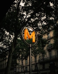 A prominent illuminated 'M' sign on a pole, likely indicating a metro or subway entrance. It is set against an urban backdrop with tall trees and an ornate early 20th-century building with balconies.