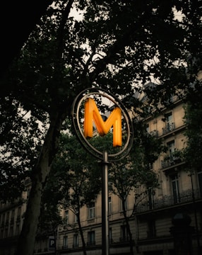 A prominent illuminated 'M' sign on a pole, likely indicating a metro or subway entrance. It is set against an urban backdrop with tall trees and an ornate early 20th-century building with balconies.