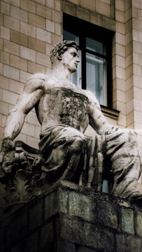A stone statue of a muscular, seated male figure is positioned against a building wall. The statue exhibits classical artistic style with detailed features and drapery over its lower body. The surrounding architecture includes rectangular stone tiles and a window, suggesting an urban or historical setting.