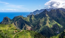 A stunning landscape features jagged mountains covered in lush greenery, extending towards the ocean. A small winding road is visible, leading to a house nestled among the hills. The sky is bright blue with scattered clouds, and the sea is a deep blue in the background.