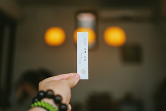 A hand is holding a small piece of paper with printed text. The background is blurred, featuring two warm-colored circular lights, resembling lamps, in a dim setting.