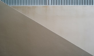 A minimalist architectural image featuring a diagonal division between two differently colored textured walls. The upper section displays a lighter shade wall with a subtle grid pattern above, while the lower section consists of a darker beige or taupe wall creating a sense of geometry and simplicity.