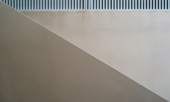 A minimalist architectural image featuring a diagonal division between two differently colored textured walls. The upper section displays a lighter shade wall with a subtle grid pattern above, while the lower section consists of a darker beige or taupe wall creating a sense of geometry and simplicity.