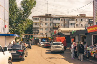 A bustling urban street scene with several parked cars and a row of small shops. People are gathered around the stores, indicating a lively market atmosphere. In the background, a large residential building rises above the scene, with many windows and balconies. Overhead, numerous electrical wires crisscross the sky.