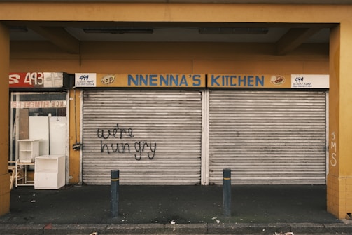 A closed storefront with metal shutters that have graffiti reading 'we're hungry'. The sign above the shutters displays 'NNENNA'S KITCHEN'. The surrounding walls are painted yellow, and there are dark posts in front of the store. A number '493' is partially visible to the side.