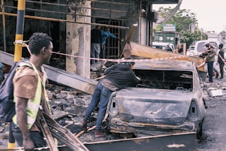 Several people are amidst the aftermath of a wrecked urban area, with debris scattered around. A burnt-out car is centrally positioned, and caution tape is draped around the site. The surroundings suggest damage from a destructive event, with individuals appearing to be assessing or involved in clean-up efforts.