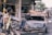 Several people are amidst the aftermath of a wrecked urban area, with debris scattered around. A burnt-out car is centrally positioned, and caution tape is draped around the site. The surroundings suggest damage from a destructive event, with individuals appearing to be assessing or involved in clean-up efforts.