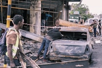 Several people are amidst the aftermath of a wrecked urban area, with debris scattered around. A burnt-out car is centrally positioned, and caution tape is draped around the site. The surroundings suggest damage from a destructive event, with individuals appearing to be assessing or involved in clean-up efforts.