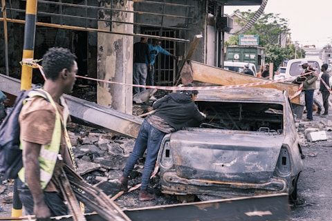 Several people are amidst the aftermath of a wrecked urban area, with debris scattered around. A burnt-out car is centrally positioned, and caution tape is draped around the site. The surroundings suggest damage from a destructive event, with individuals appearing to be assessing or involved in clean-up efforts.