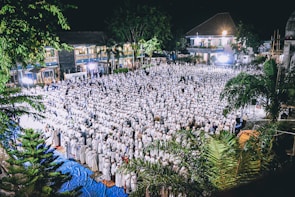 A large crowd of people dressed in white is gathered for a community event at night, likely a religious or cultural gathering. The setting includes outdoor space with surrounding trees and buildings, illuminated by bright lights.