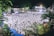 A large crowd of people dressed in white is gathered for a community event at night, likely a religious or cultural gathering. The setting includes outdoor space with surrounding trees and buildings, illuminated by bright lights.