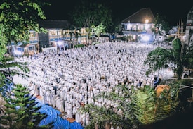 A large crowd of people dressed in white is gathered for a community event at night, likely a religious or cultural gathering. The setting includes outdoor space with surrounding trees and buildings, illuminated by bright lights.
