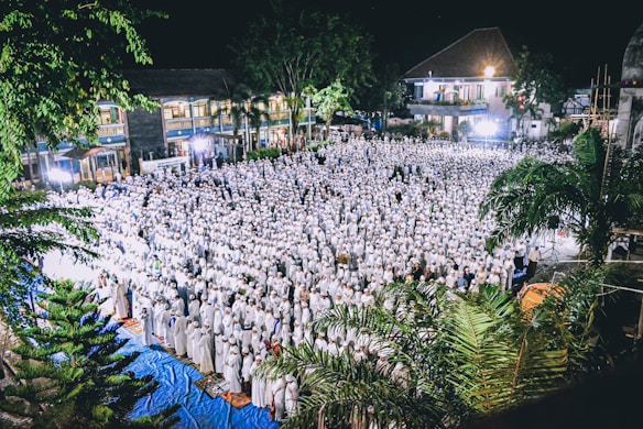 A large crowd of people dressed in white is gathered for a community event at night, likely a religious or cultural gathering. The setting includes outdoor space with surrounding trees and buildings, illuminated by bright lights.