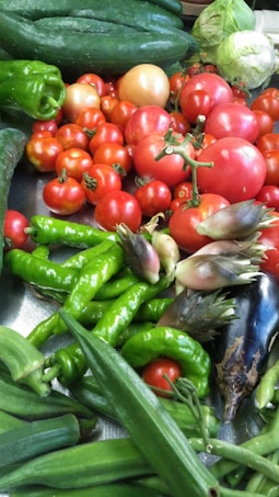 A vibrant assortment of fresh vegetables, including bright red tomatoes, green peppers, cucumbers, okra, eggplants, and cabbage. The vegetables are piled together, showcasing a variety of shapes and sizes, with a glossy surface on some of them reflecting light.