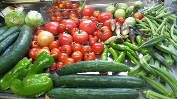 A collection of various fresh vegetables is arranged on a surface. There are ripe tomatoes, cucumbers, lettuce, green bell peppers, okra, and other assorted produce.