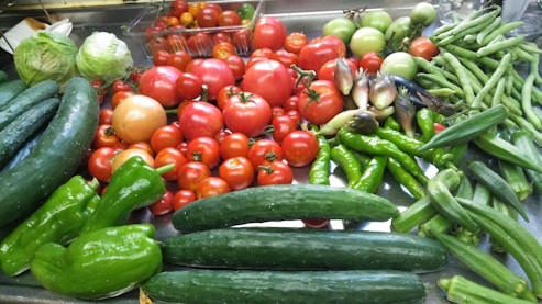 A collection of various fresh vegetables is arranged on a surface. There are ripe tomatoes, cucumbers, lettuce, green bell peppers, okra, and other assorted produce.