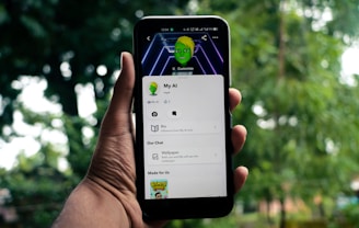 A hand holds a smartphone displaying an app screen with cartoonish graphics featuring a green-skinned avatar. The app interface includes options like 'Bio' and 'Wallpaper'. The background is blurred, showcasing greenery.