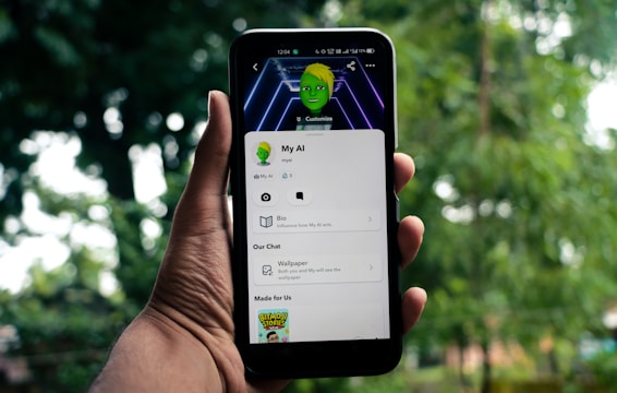 A hand holds a smartphone displaying an app screen with cartoonish graphics featuring a green-skinned avatar. The app interface includes options like 'Bio' and 'Wallpaper'. The background is blurred, showcasing greenery.