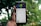 A hand holds a smartphone displaying an app screen with cartoonish graphics featuring a green-skinned avatar. The app interface includes options like 'Bio' and 'Wallpaper'. The background is blurred, showcasing greenery.