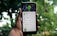 A hand holds a smartphone displaying an app screen with cartoonish graphics featuring a green-skinned avatar. The app interface includes options like 'Bio' and 'Wallpaper'. The background is blurred, showcasing greenery.