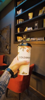 A hand with a tattoo holds a cup labeled 'Cold Brew' in front of a cozy cafe setting. The background features a wall with wooden bookshelves filled with books and decorative items, including a cup and a 'Reserved' sign. A red chair is visible beside a dark-colored table.