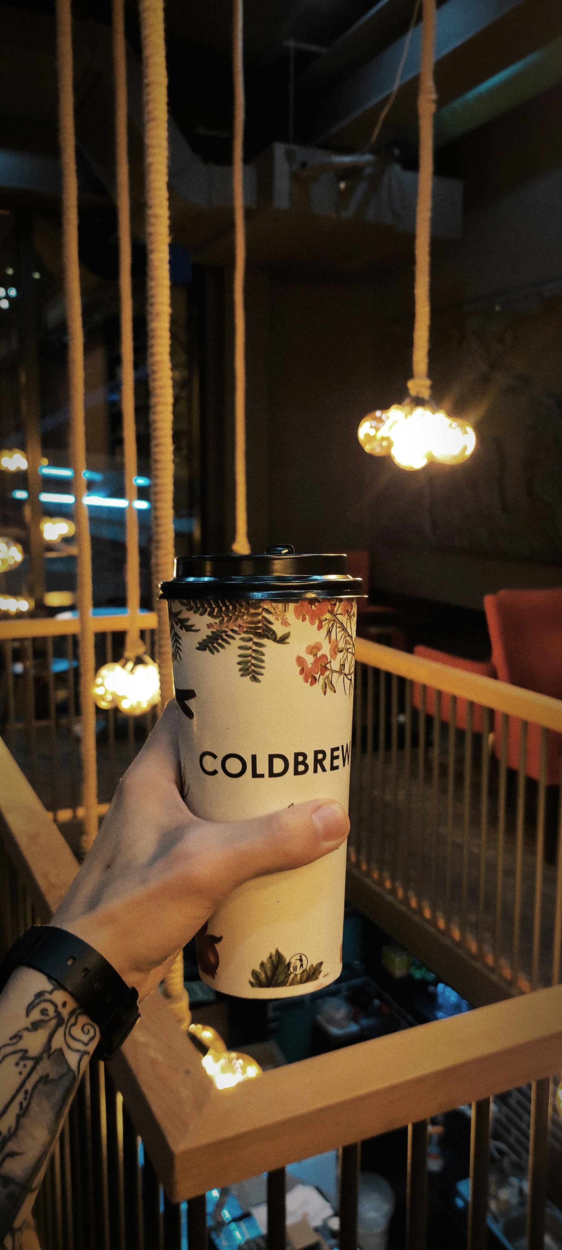 A person with a tattooed arm holds up a cup labeled 'COLDBREW' in a dimly lit cafe. The cafe features warm lighting with hanging bulbs and wooden railings, creating a cozy ambiance. A red chair is visible in the background.
