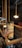 A person with a tattooed arm holds up a cup labeled 'COLDBREW' in a dimly lit cafe. The cafe features warm lighting with hanging bulbs and wooden railings, creating a cozy ambiance. A red chair is visible in the background.