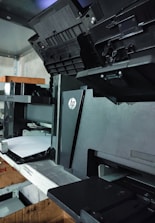 A black HP printer is positioned on a metal shelf in a storage area. The printer has opened compartments and trays, with an additional tray containing some white paper loaded into it. The surroundings include other electronic devices and boxes on the same and nearby shelves.