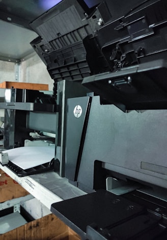 A black HP printer is positioned on a metal shelf in a storage area. The printer has opened compartments and trays, with an additional tray containing some white paper loaded into it. The surroundings include other electronic devices and boxes on the same and nearby shelves.