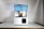 A 3D printer with a blue translucent cover is placed on a reflective surface. It has a rectangular base and an open chamber where the printing mechanism is visible. The power cord extends from the back and it is set against a plain background.