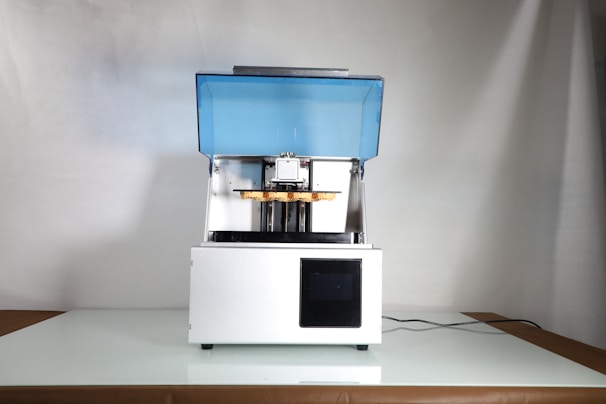 A 3D printer with a blue translucent cover is placed on a reflective surface. It has a rectangular base and an open chamber where the printing mechanism is visible. The power cord extends from the back and it is set against a plain background.