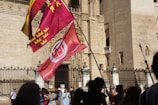 A group of people standing in front of an ornate, historic building with a large gate. Several colorful flags, including red and yellow ones, are prominently displayed, blowing in the breeze. The architectural details of the building suggest it is of significant cultural or historic value. Shadows of the people can be seen on the stone facade, indicating bright sunlight.