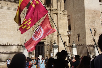 A group of people standing in front of an ornate, historic building with a large gate. Several colorful flags, including red and yellow ones, are prominently displayed, blowing in the breeze. The architectural details of the building suggest it is of significant cultural or historic value. Shadows of the people can be seen on the stone facade, indicating bright sunlight.