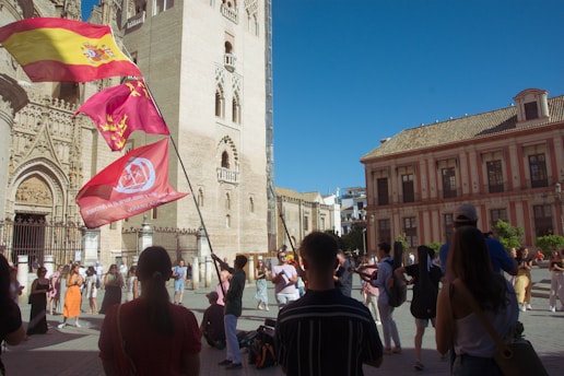 A lively street scene with a crowd gathered near a historical building, likely a cathedral, featuring intricate architectural details. Multiple flags are being waved, one of which is the Spanish flag. People appear to be engaged in an organized event or celebration.