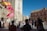 A lively street scene with a crowd gathered near a historical building, likely a cathedral, featuring intricate architectural details. Multiple flags are being waved, one of which is the Spanish flag. People appear to be engaged in an organized event or celebration.